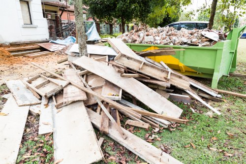 A pile of discarded wooden decking boards and broken timber planks with rough, weathered textures and light brown and grey tones, resting on a grassy area adjacent to a small, green industrial skip filled with mixed rubble and bricks, in an outdoor setting near residential buildings and trees, with some debris scattered around and a background of a pavement and garden space typical of a Wembley suburb. The scene appears to be part of an ongoing garden clearance or rubbish removal process coordinated by Garden Clearance Wembley to clear out construction and garden waste, emphasizing the professional handling of rubbish collection in the local area.