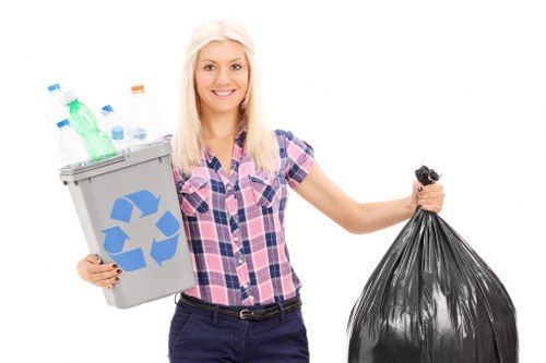 A young woman with long blonde hair, dressed in a casual pink and blue checked shirt and dark trousers, stands against a plain white background. She is holding a small, grey recycling bin filled with various used plastic bottles, including green, clear, and blue bottles, with some caps visible. In her other hand, she holds a large, black plastic rubbish bag that appears to be filled with waste materials. Her expression is friendly, with a slight smile. The scene is neutral and composed to illustrate waste management or rubbish disposal, consistent with services offered by Garden Clearance Wembley. Although the background is plain, the focus is on the woman and the waste items she holds, representing rubbish removal and recycling efforts typical in professional waste clearance in Wembley and surrounding areas.