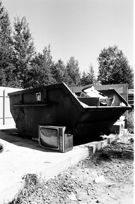 A large, black, metal skip bin with a rectangular shape, situated outdoors on a concrete surface. The bin is partially filled and appears weathered, with visible rust and dirt marks on its exterior. I
