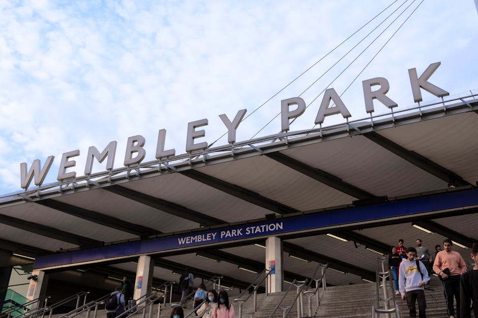 An exterior view of Wembley Stadium during dusk, with the stadium's curved metal roof structure visible at the top left corner, featuring an intricate lattice framework. Below, a large turquoise sign