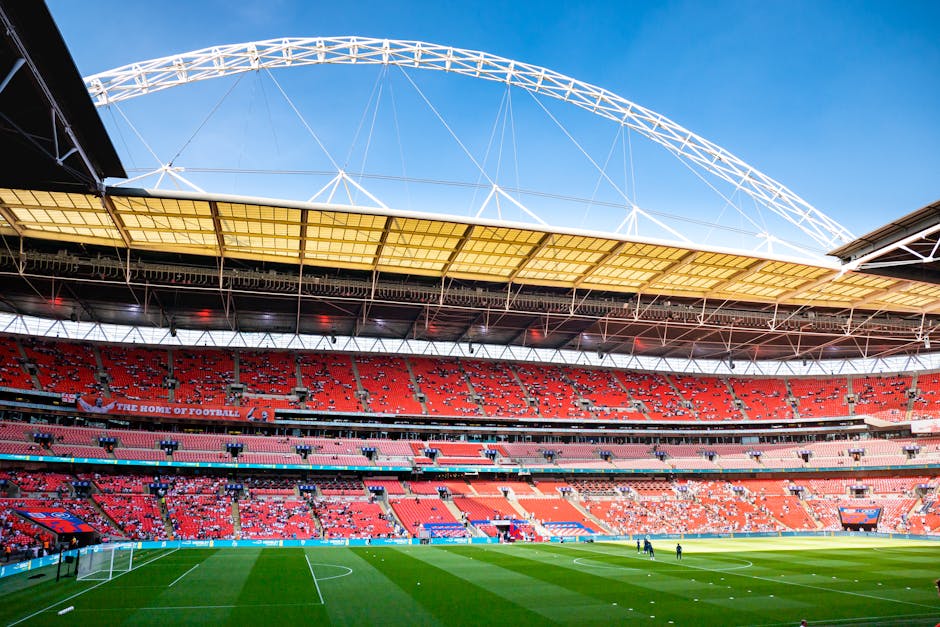 A panoramic view of Wembley Stadium with its distinctive white arched roof structure set against a clear blue sky. The seating areas are predominantly red, with sections of empty seats visible and som