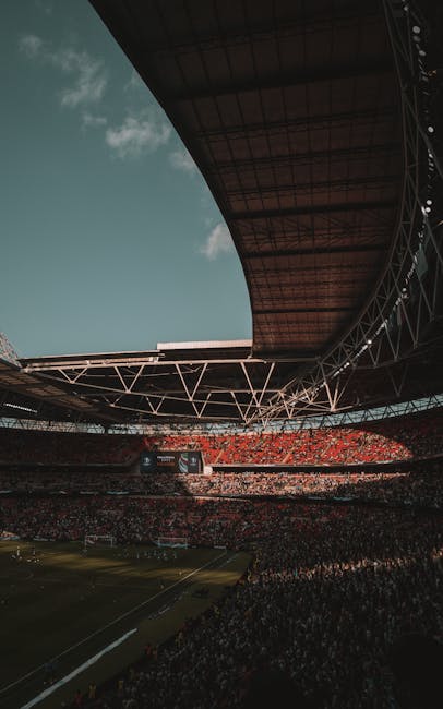 A panoramic view of Wembley Stadium with its distinctive white arched roof structure set against a clear blue sky. The seating areas are predominantly red, with sections of empty seats visible and som